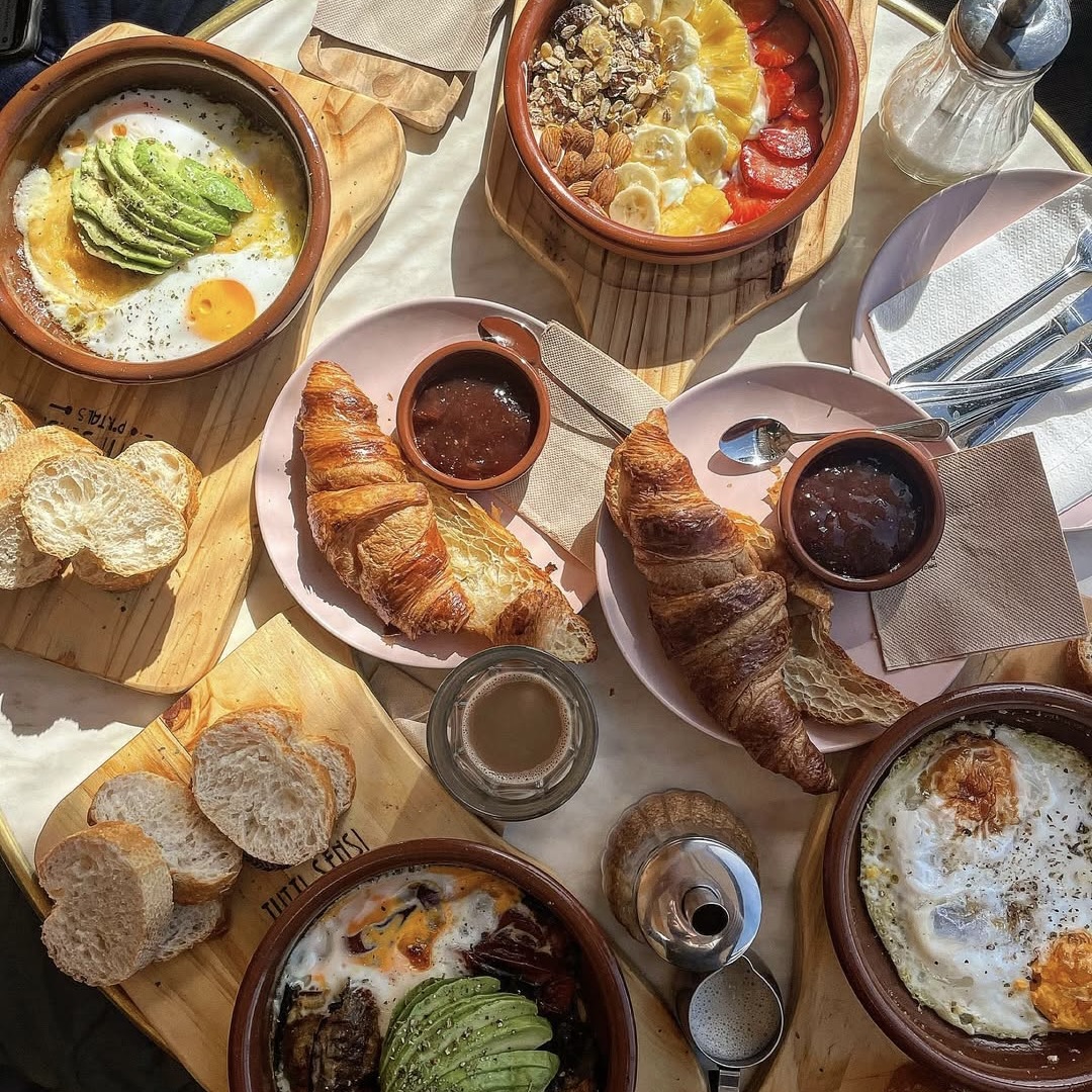 Desayuno italiano frente al mar en Puerto Portals con croissants, aguacate, bol de fruta, pan, huevos y café, restaurante con terraza exterior e interior.